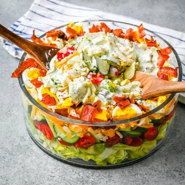 This classic Seven Layer salad is layered with iceberg lettuce, smoky bacon, hard-boiled eggs, sharp cheddar cheese, fresh veggies, and your choice of peas or garbanzo beans, plus creamy avocado. It's the ultimate make-ahead side dish, perfect for BBQs or family dinners. This easy, crowd-pleasing 7 Layer Salad is topped with a generous drizzle of homemade ranch dressing. This easy, crowd-pleasing layered salad is a beautiful, delicious side dish!