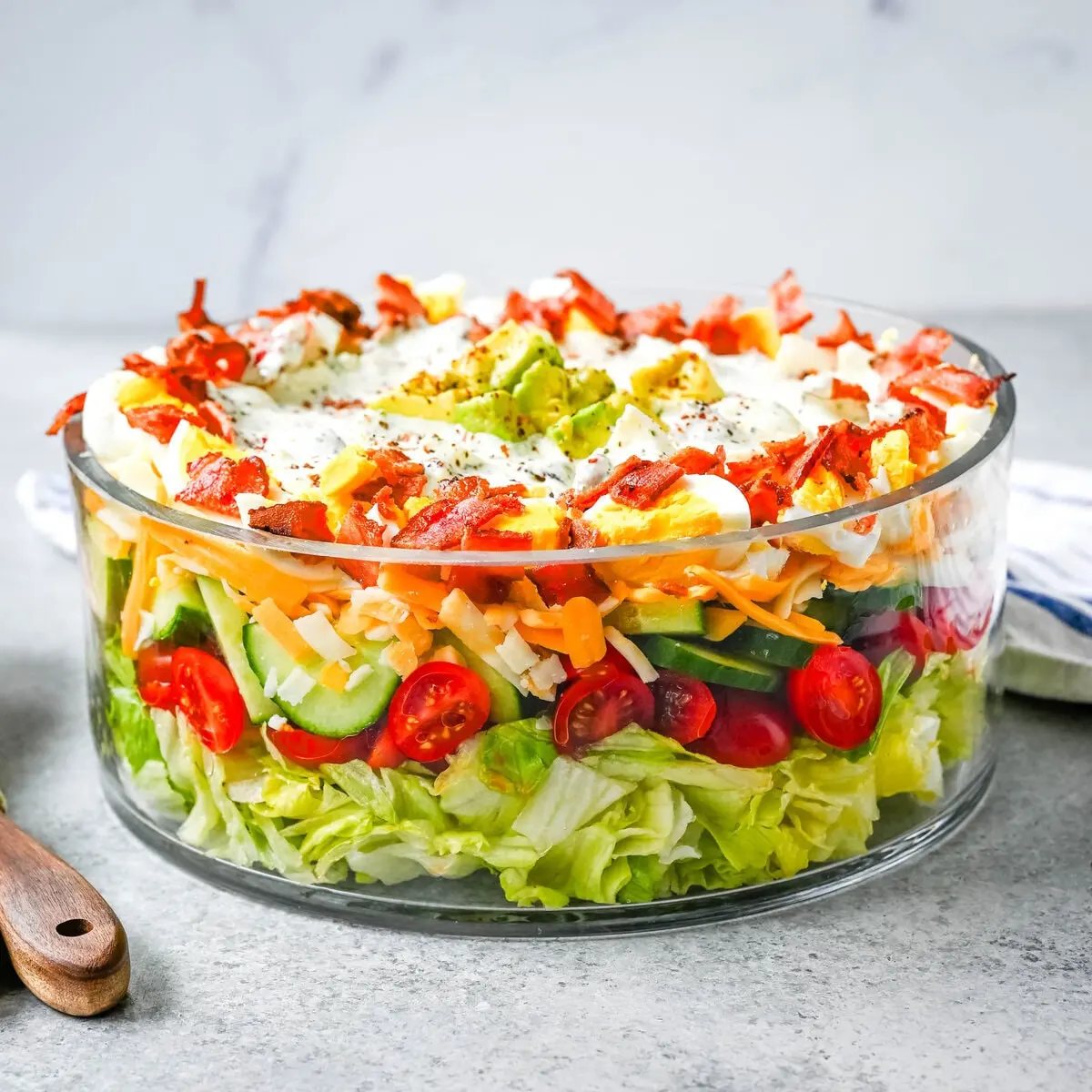 This classic Seven Layer salad is layered with iceberg lettuce, smoky bacon, hard-boiled eggs, sharp cheddar cheese, fresh veggies, and your choice of peas or garbanzo beans, plus creamy avocado. It's the ultimate make-ahead side dish, perfect for BBQs or family dinners. This easy, crowd-pleasing 7 Layer Salad is topped with a generous drizzle of homemade ranch dressing. This easy, crowd-pleasing layered salad is a beautiful, delicious side dish!