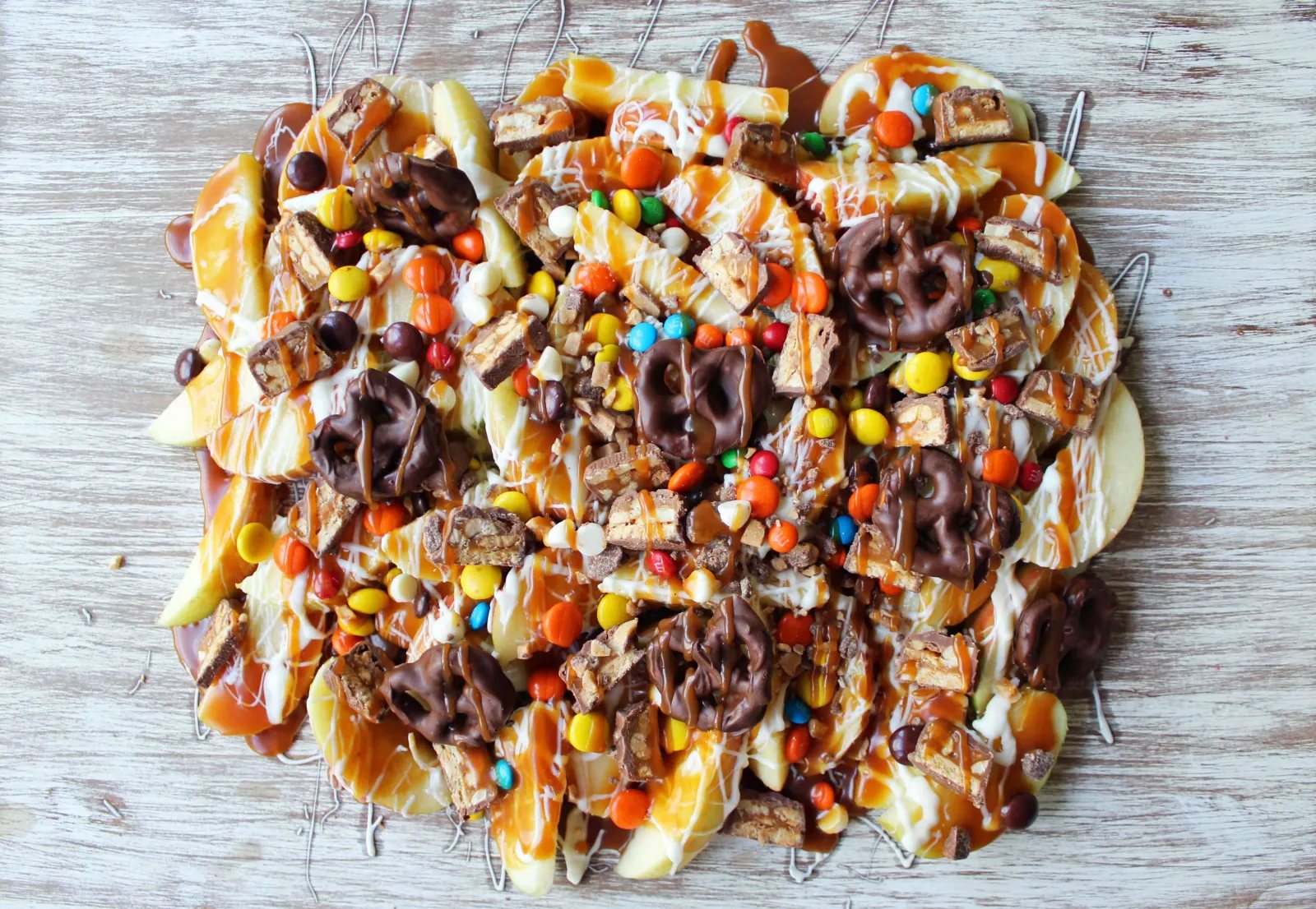 Candy Madness Caramel Apple Nachos by Modern Honey. Crisp Apples topped with Salted Caramel, White Chocolate, M &M's, Reeses Pieces, Heath Toffee Bits, Snickers, and Chocolate Covered Pretzels. These Caramel Apple Nachos are much easier than making caramel apples and will be a huge hit at your next party!