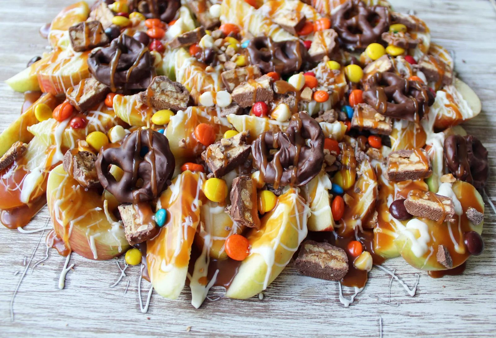 Candy Madness Caramel Apple Nachos by Modern Honey. Crisp Apples topped with Salted Caramel, White Chocolate, M &M's, Reeses Pieces, Heath Toffee Bits, Snickers, and Chocolate Covered Pretzels. These Caramel Apple Nachos are much easier than making caramel apples and will be a huge hit at your next party!