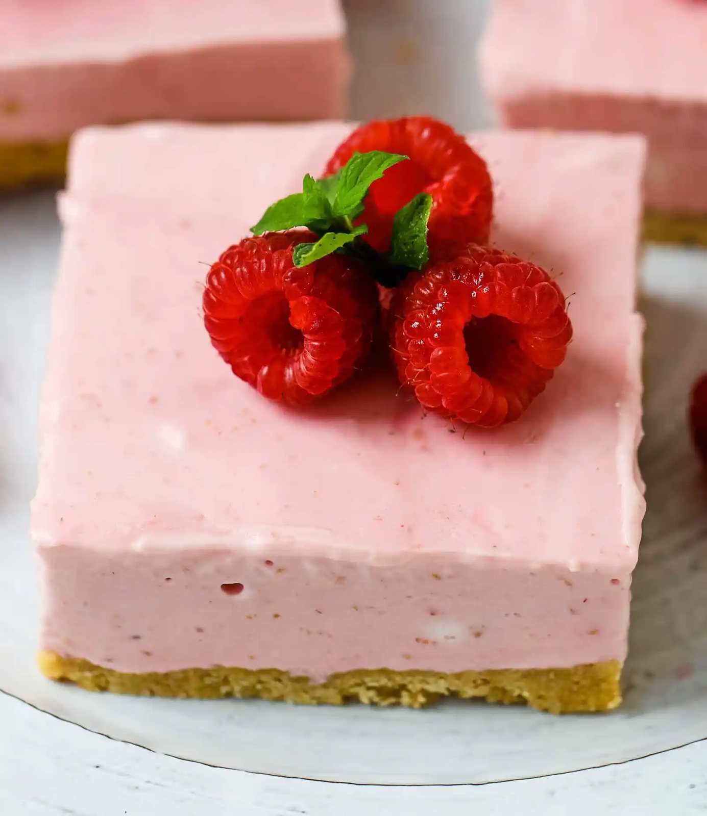 No-Bake Frozen Raspberry Cheesecake Squares. Creamy no-bake raspberry cheesecake filling on top of a shortbread or graham cracker crust topped with fresh raspberries. A creamy no-bake frozen dessert. www.modernhoney.com #nobakedessert #raspberrycheesecake #cheesecakesquares #frozendessert #frozencheesecake