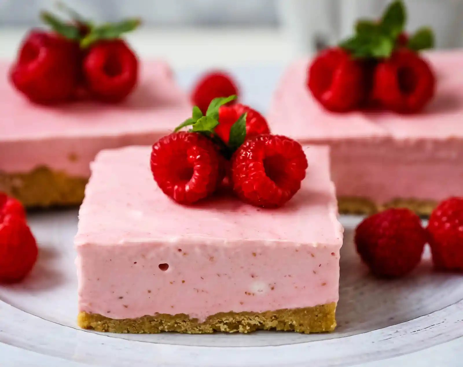 No-Bake Frozen Raspberry Cheesecake Squares. Creamy no-bake raspberry cheesecake filling on top of a shortbread or graham cracker crust topped with fresh raspberries. A creamy no-bake frozen dessert. www.modernhoney.com #nobakedessert #raspberrycheesecake #cheesecakesquares #frozendessert #frozencheesecake