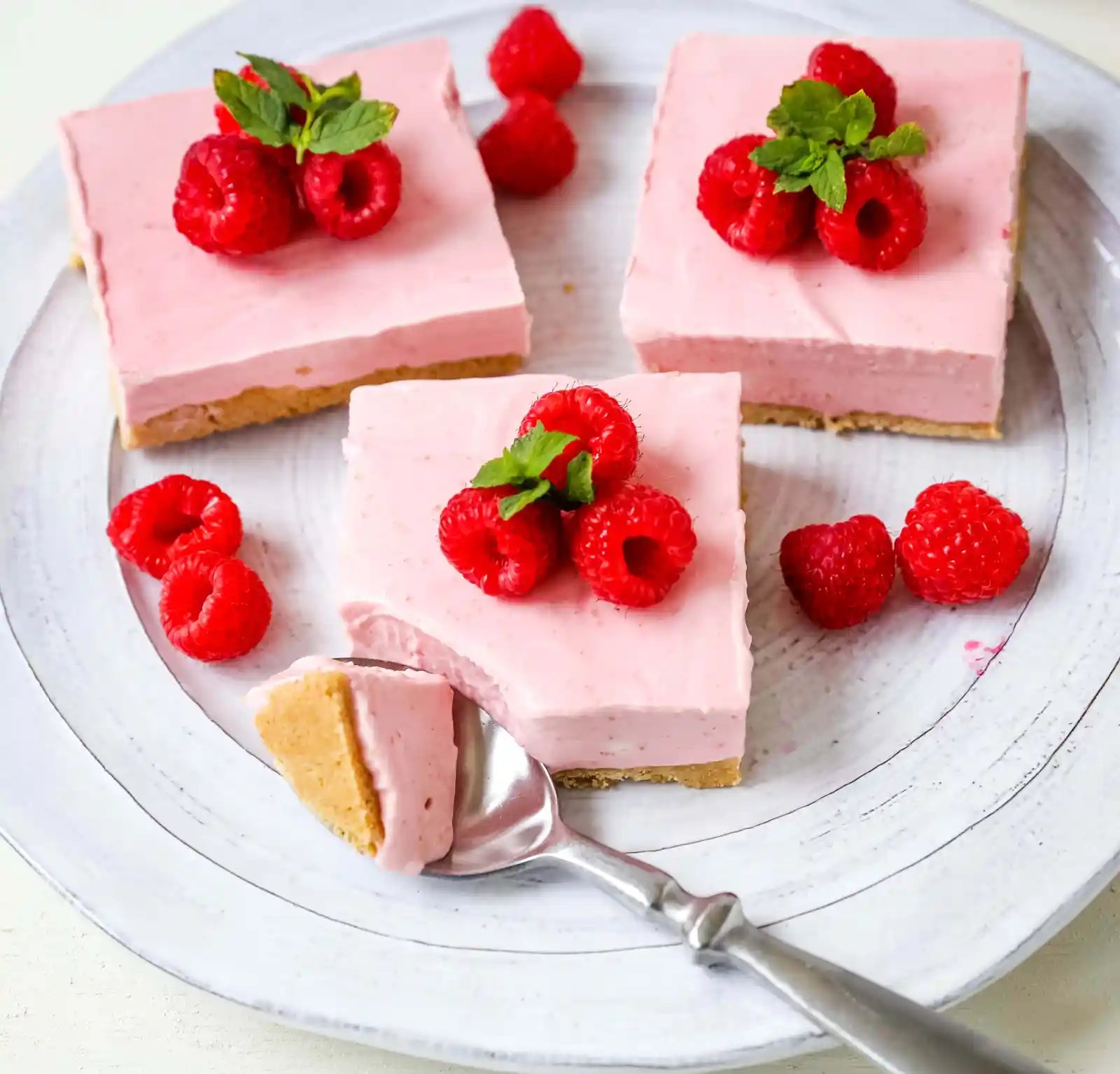 No-Bake Frozen Raspberry Cheesecake Squares. Creamy no-bake raspberry cheesecake filling on top of a shortbread or graham cracker crust topped with fresh raspberries. A creamy no-bake frozen dessert. www.modernhoney.com #nobakedessert #raspberrycheesecake #cheesecakesquares #frozendessert #frozencheesecake