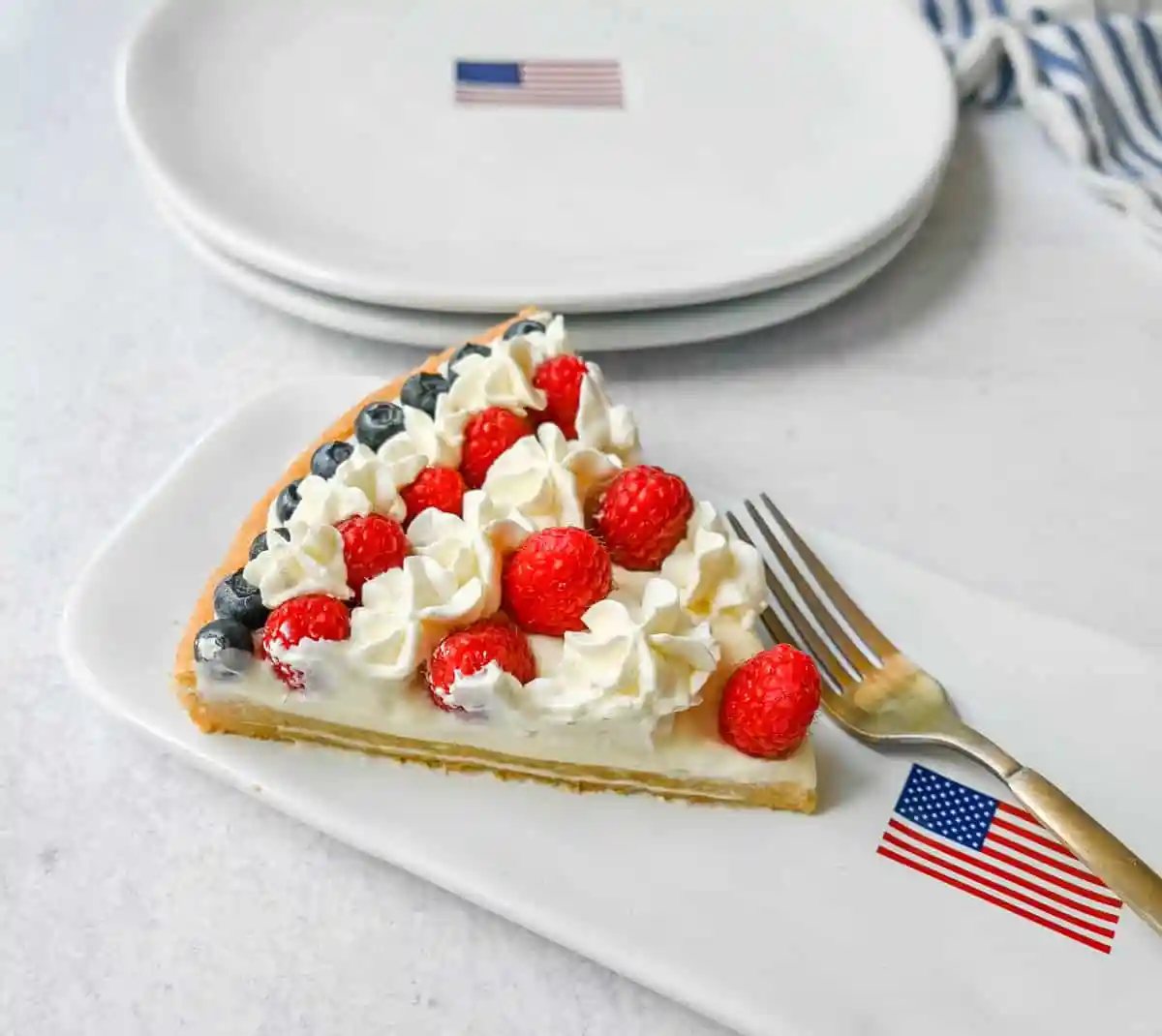 This flag sugar cookie fruit pizza is a festive, delicious, and beautiful patriotic 4th of July dessert featuring a homemade soft sugar cookie crust with a sweet vanilla cream cheese frosting topped with fresh raspberries, blueberries, and homemade whipped cream.