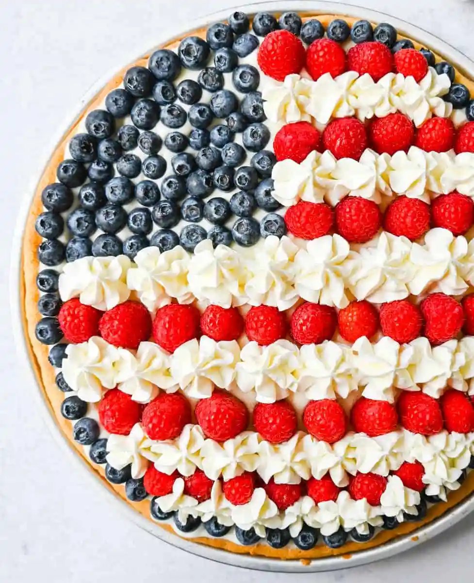 This flag sugar cookie fruit pizza is a festive, delicious, and beautiful patriotic 4th of July dessert featuring a homemade soft sugar cookie crust with a sweet vanilla cream cheese frosting topped with fresh raspberries, blueberries, and homemade whipped cream.