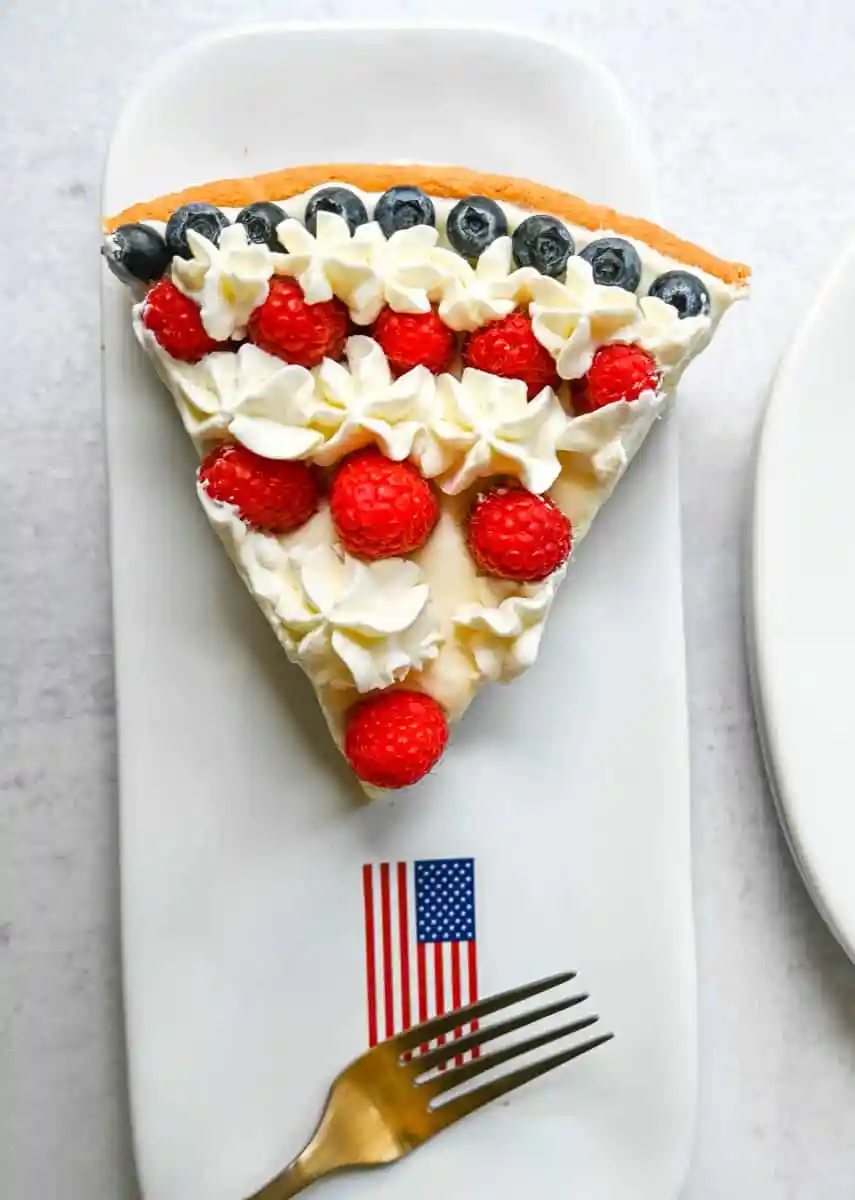 This flag sugar cookie fruit pizza is a festive, delicious, and beautiful patriotic 4th of July dessert featuring a homemade soft sugar cookie crust with a sweet vanilla cream cheese frosting topped with fresh raspberries, blueberries, and homemade whipped cream.