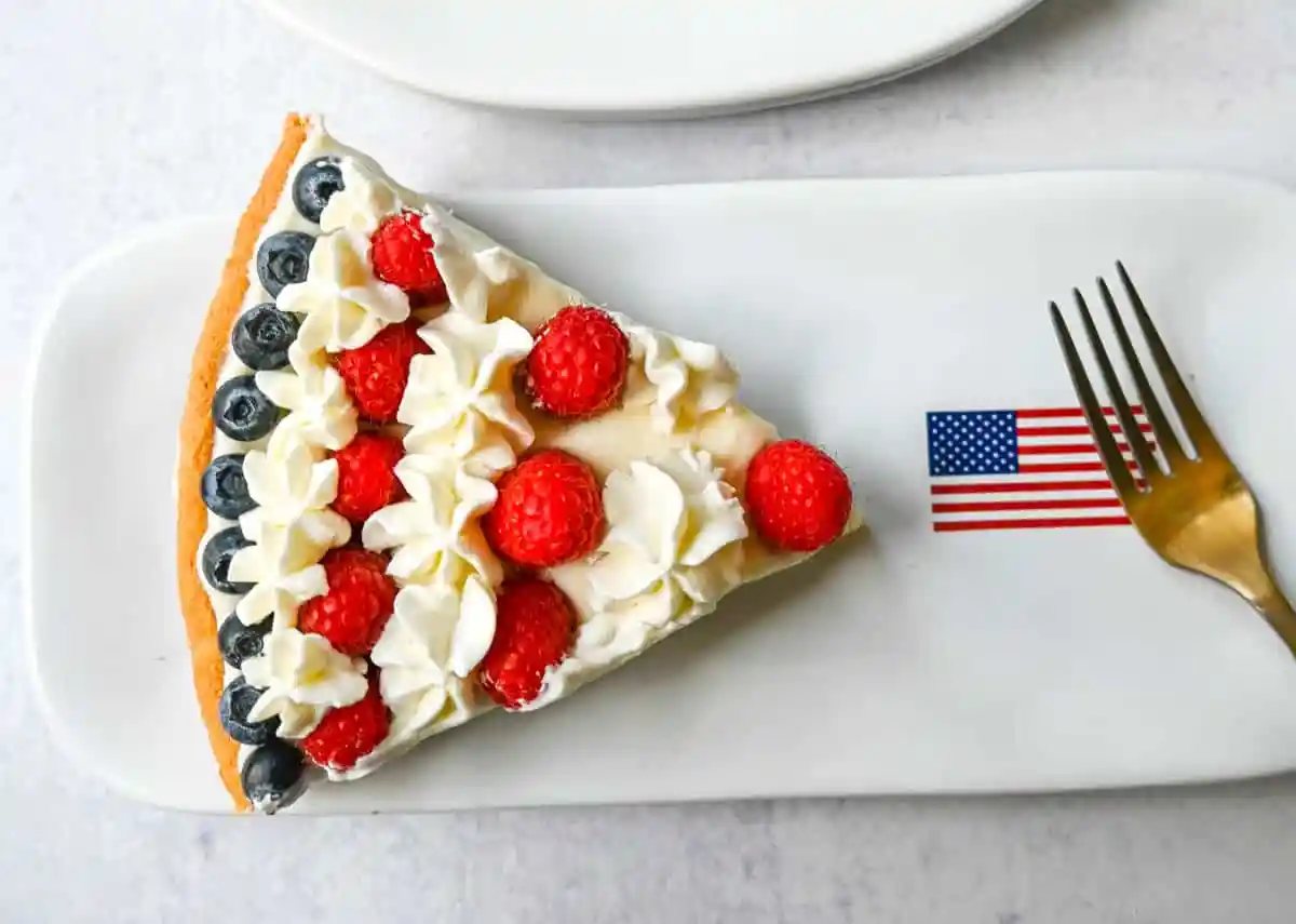 This flag sugar cookie fruit pizza is a festive, delicious, and beautiful patriotic 4th of July dessert featuring a homemade soft sugar cookie crust with a sweet vanilla cream cheese frosting topped with fresh raspberries, blueberries, and homemade whipped cream.