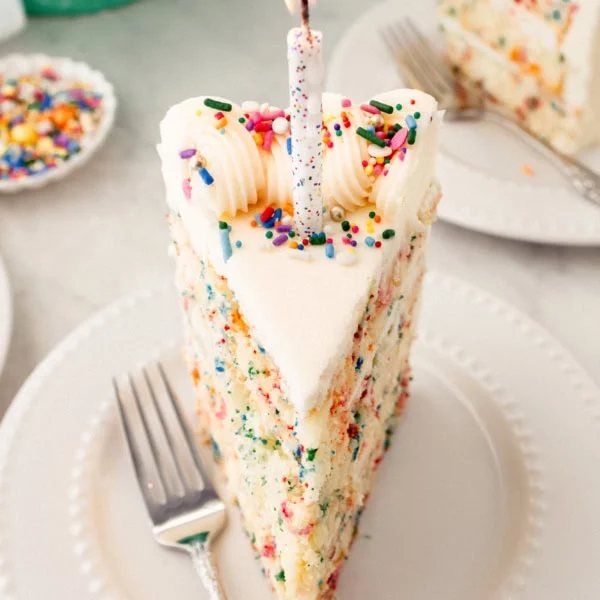 Piece of birthday cake with sprinkles with a candle. A fluffy homemade Funfetti birthday cake with rainbow sprinkles topped with a light cream cheese buttercream frosting and an array of sprinkles. This Confetti Cake recipe is the perfect birthday cake!