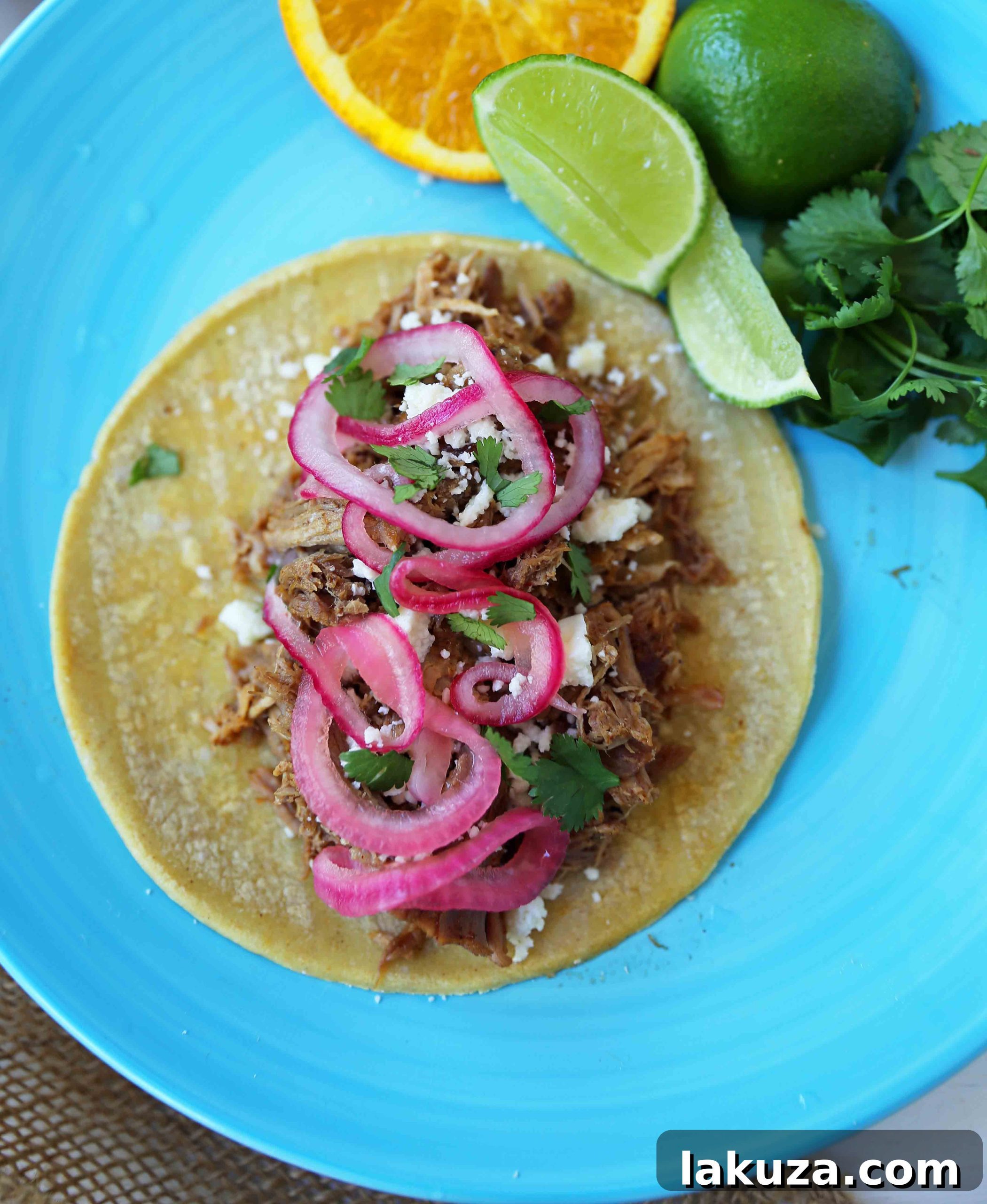 A vibrant close-up of shredded Mexican Pork Carnitas, beautifully crisped, served with a generous garnish of bright pickled red onions, fresh cilantro, and a lime wedge, ready to be assembled into delicious tacos.