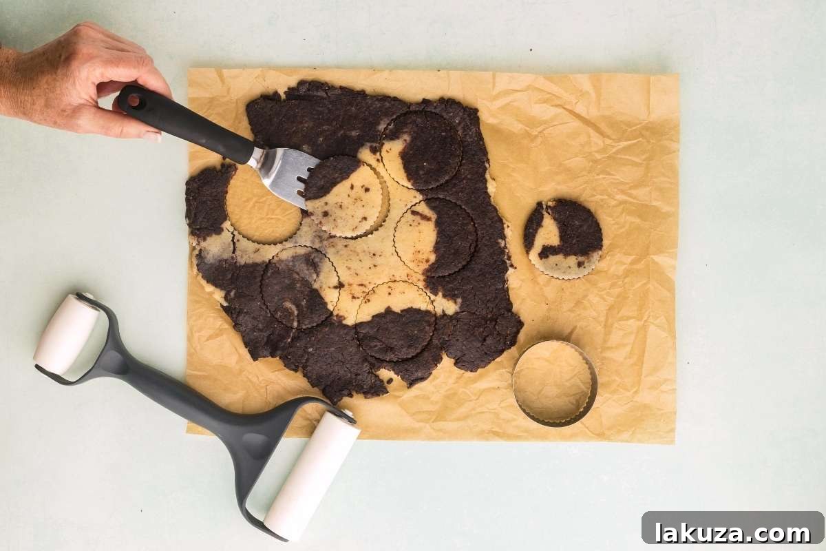 Hands using a round cookie cutter to cut out cookies from the swirled shortbread dough on parchment paper.