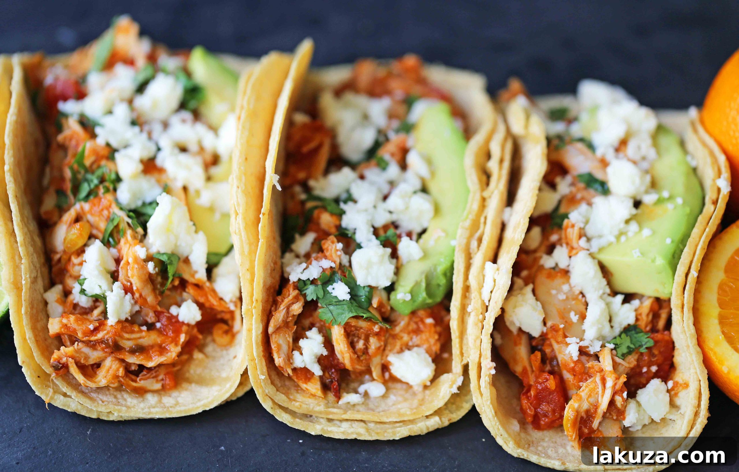 Close-up of three Chipotle Chicken Tinga Tacos on a wooden board, garnished with cotija cheese, cilantro, and avocado slices. A simple, quick, and easy chicken taco recipe, ideal for a one-skillet preparation.