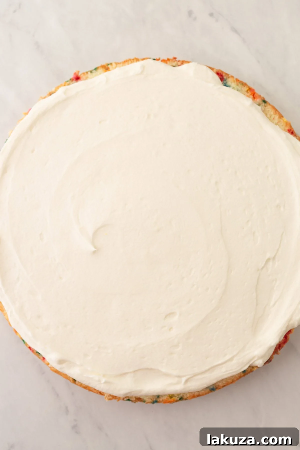 Joyful Sprinkle Celebration 12 A skilled hand using an offset spatula to apply smooth vanilla cream cheese frosting onto a layer of homemade Funfetti cake, preparing it for assembly.