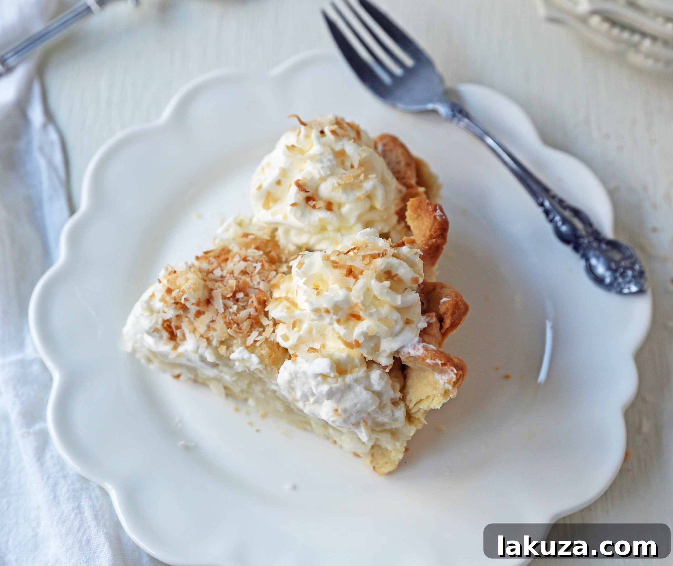 A beautifully presented whole Coconut Cream Pie, decorated with elegant swirls of whipped cream and toasted coconut, on a white cake stand.