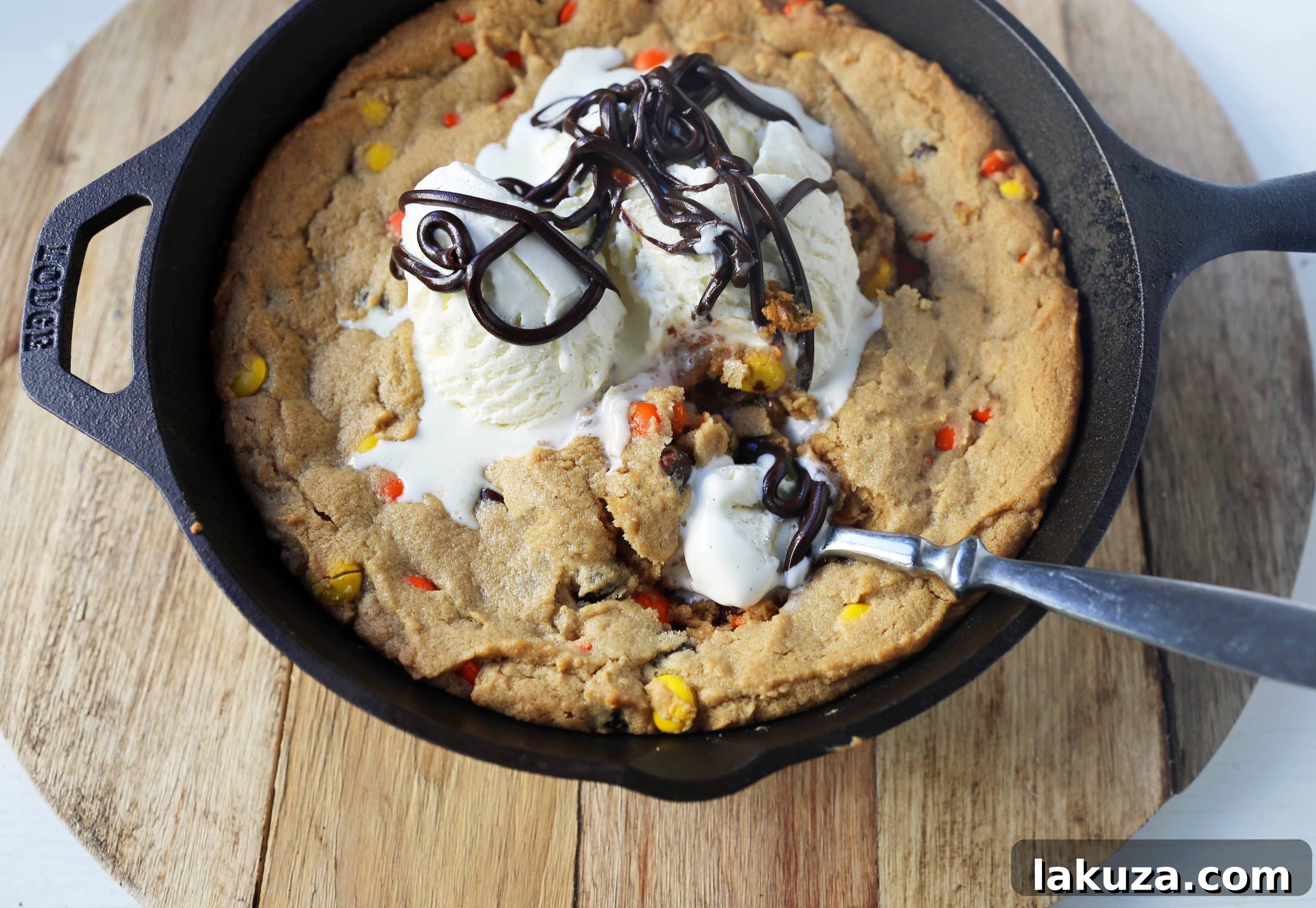 Close-up of a warm, deep-dish Peanut Butter Skillet Cookie filled with melted Reese's candies, served with a scoop of vanilla ice cream and chocolate syrup.