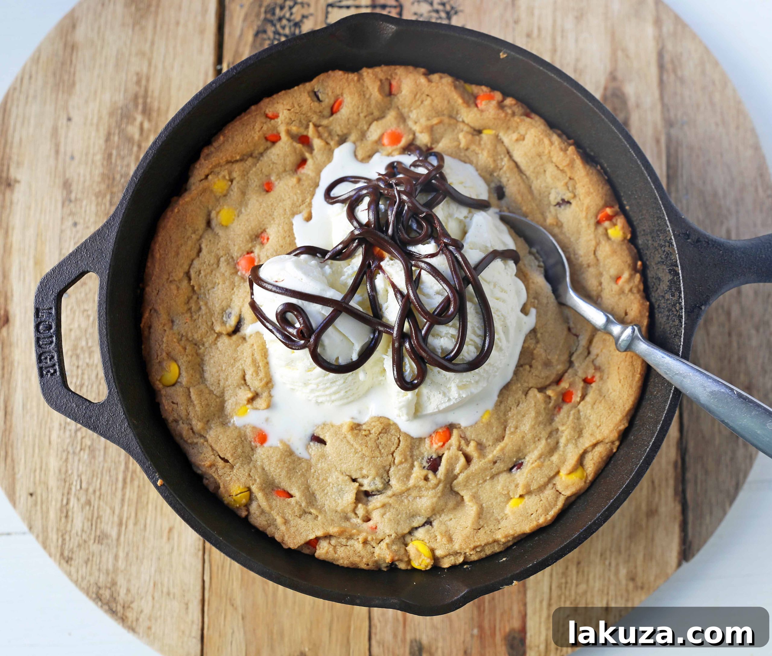 Hand scooping vanilla ice cream onto a freshly baked Peanut Butter Skillet Cookie in a cast iron pan, ready to be drizzled with hot fudge.