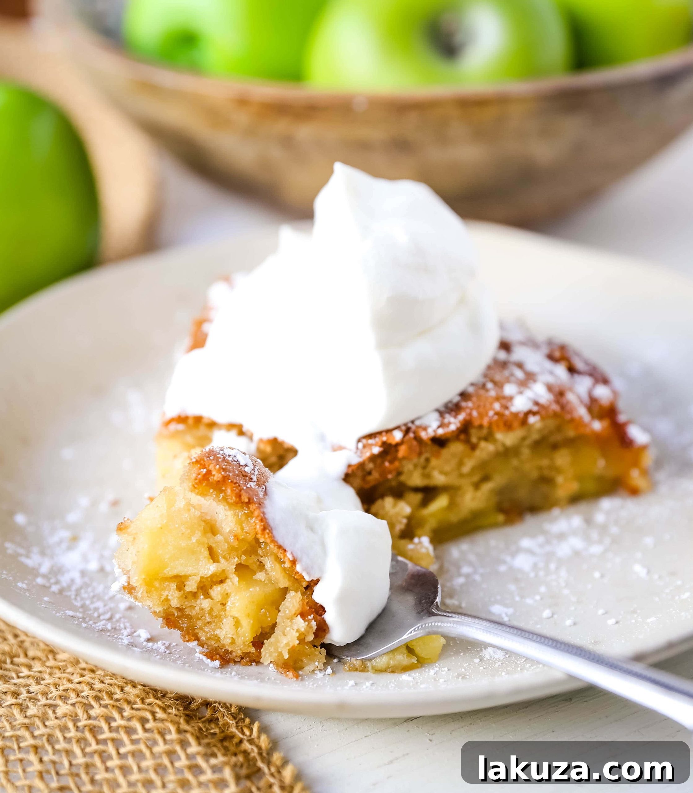 A close-up shot of French Apple Cake slices, generously dusted with powdered sugar and adorned with fresh whipped cream. The golden crust and tender apples promise a delightful dessert.