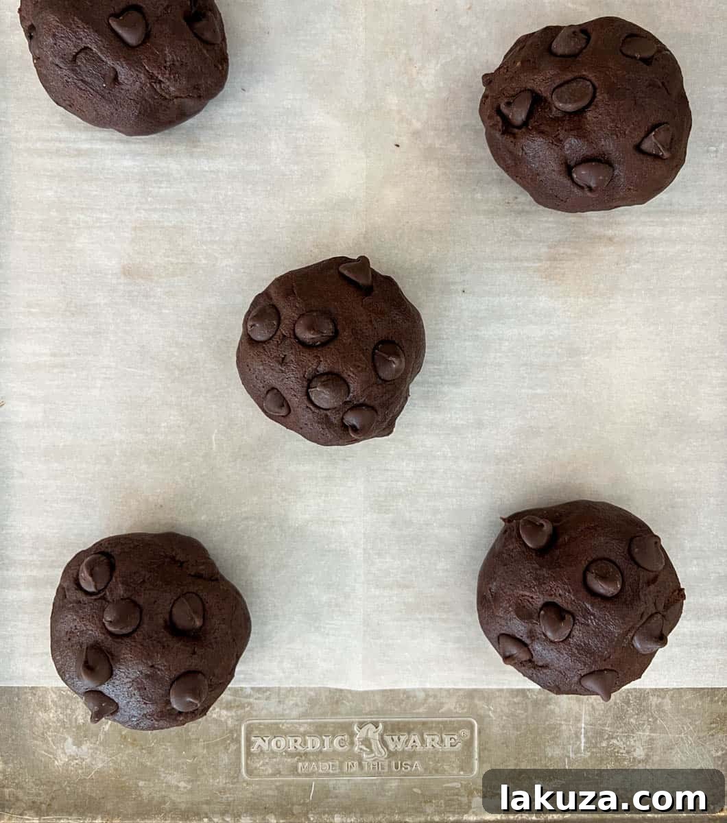 Portioned balls of double chocolate chip cookie dough arranged on a baking sheet lined with parchment paper, some with extra chocolate chips pressed into the surface for a gourmet look.