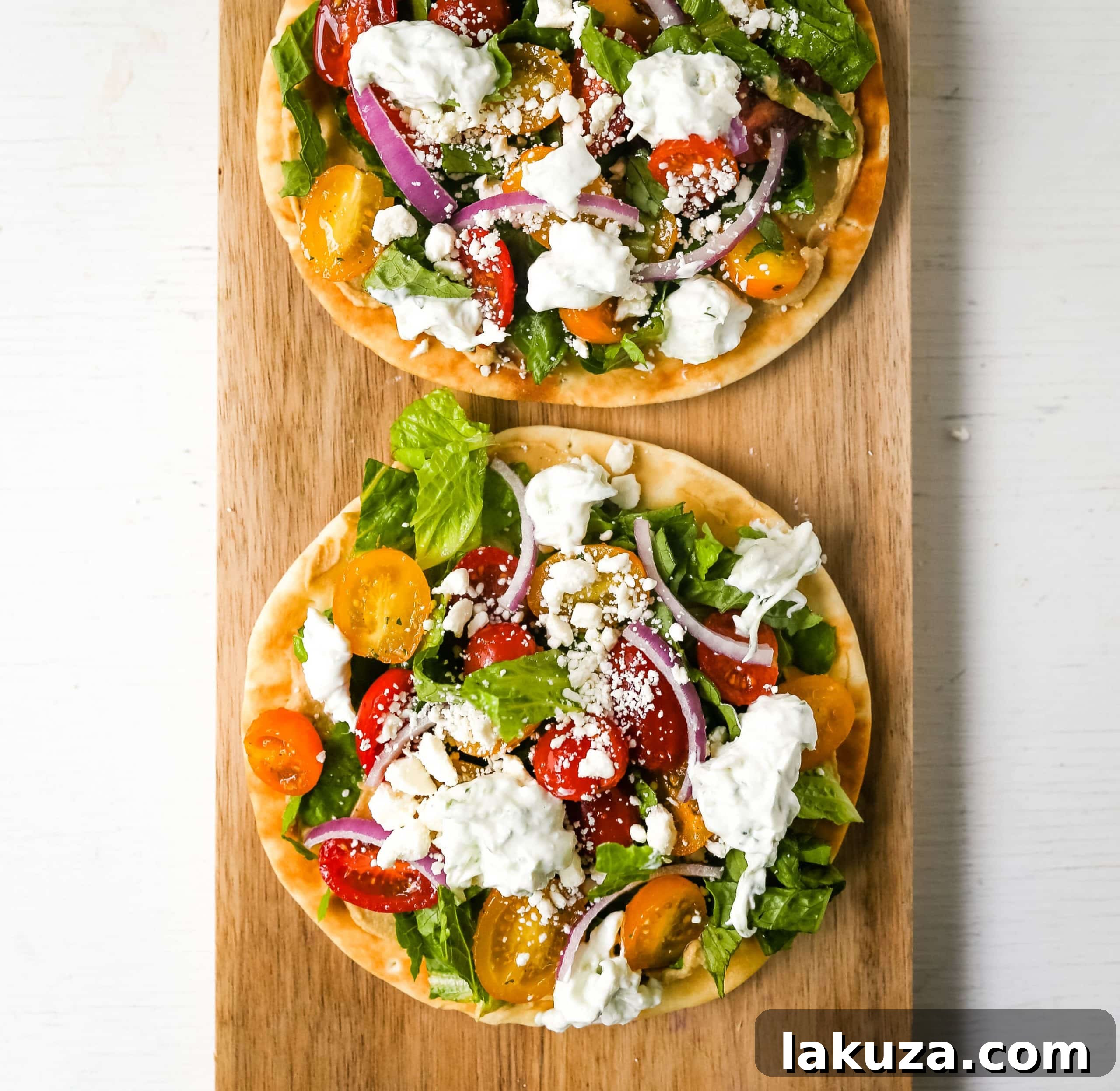 Zesty Greek Pita Pizzas 3 Close-up of an individual Greek Salad Pita Pizza, showcasing the layers of creamy hummus on soft pita, topped with vibrant green lettuce, halved red and yellow grape tomatoes, thin cucumber slices, red onion, and white feta cheese, ready to be drizzled with tzatziki.
