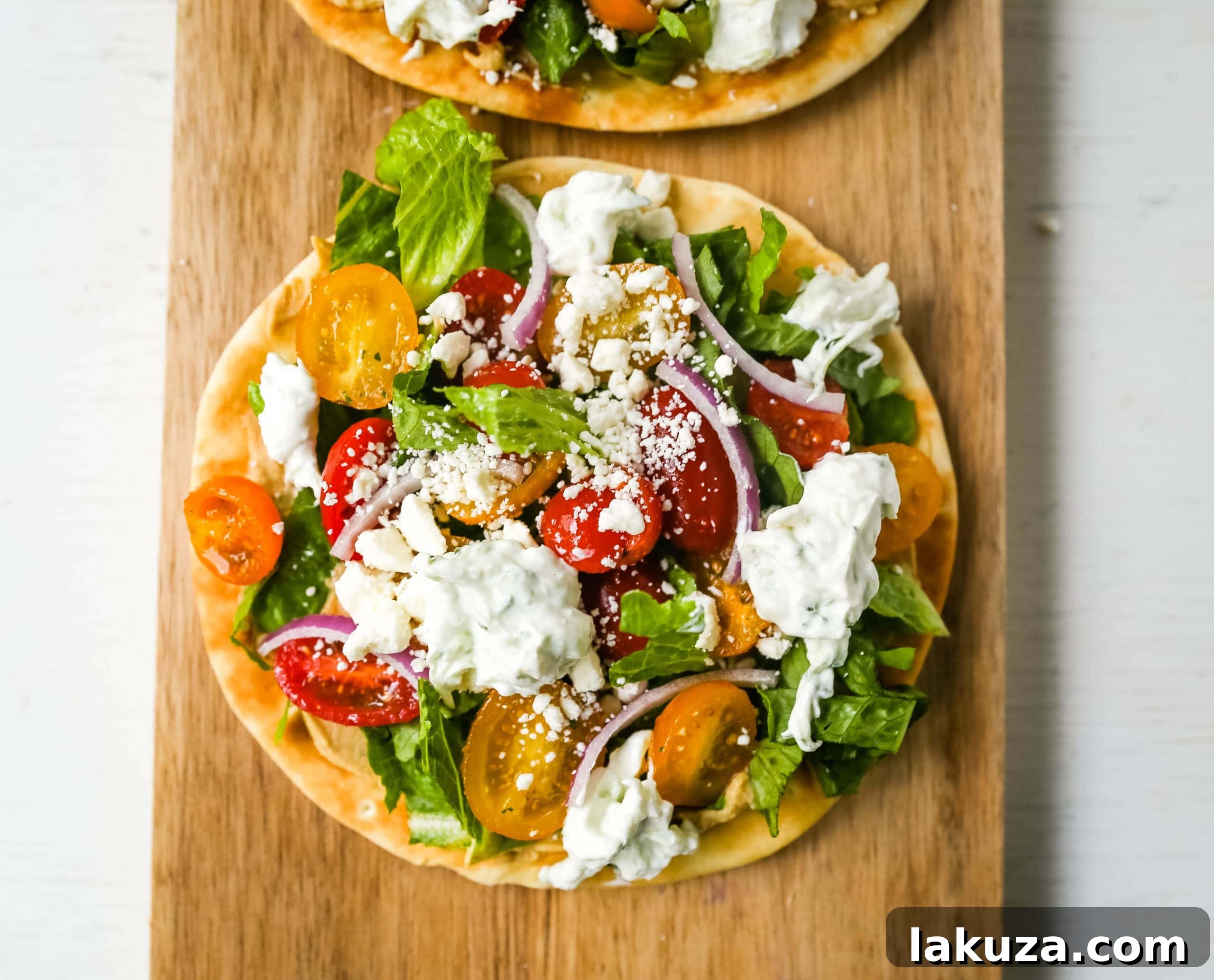 Zesty Greek Pita Pizzas 4 Three perfectly warmed pita bread bases, each spread with a thick layer of creamy hummus, waiting for their fresh Greek salad toppings, with colorful grape tomatoes visible in a bowl nearby.