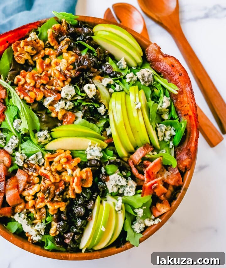 Overhead view of a prepared Fall Harvest Salad with all components mixed and ready to be dressed: mixed greens, apple slices, candied walnuts, dried cranberries, crumbled cheese, and crispy bacon.