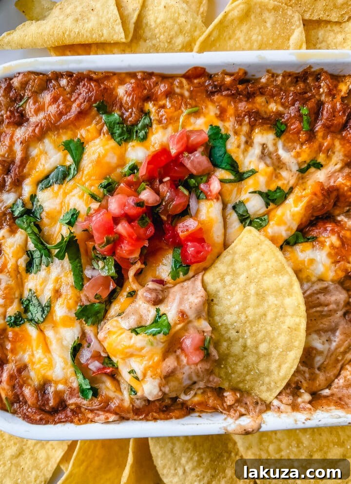 A large serving of hot bean and cheese dip in a baking dish, with a scoop taken out, revealing the creamy layers. It's surrounded by tortilla chips.