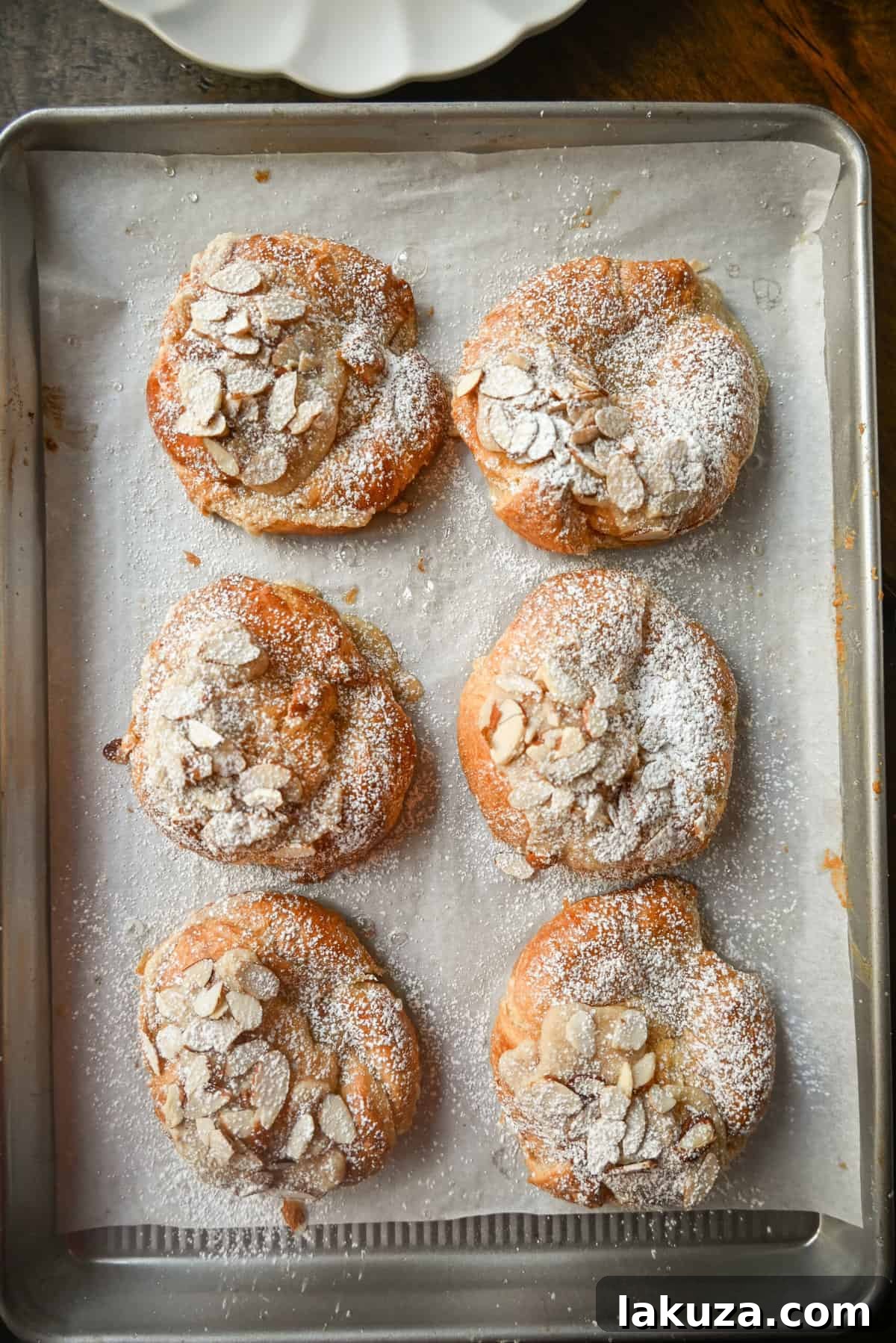Raw almond croissants on a baking sheet, brushed with syrup, filled with frangipane, and topped with sliced almonds, ready for baking.