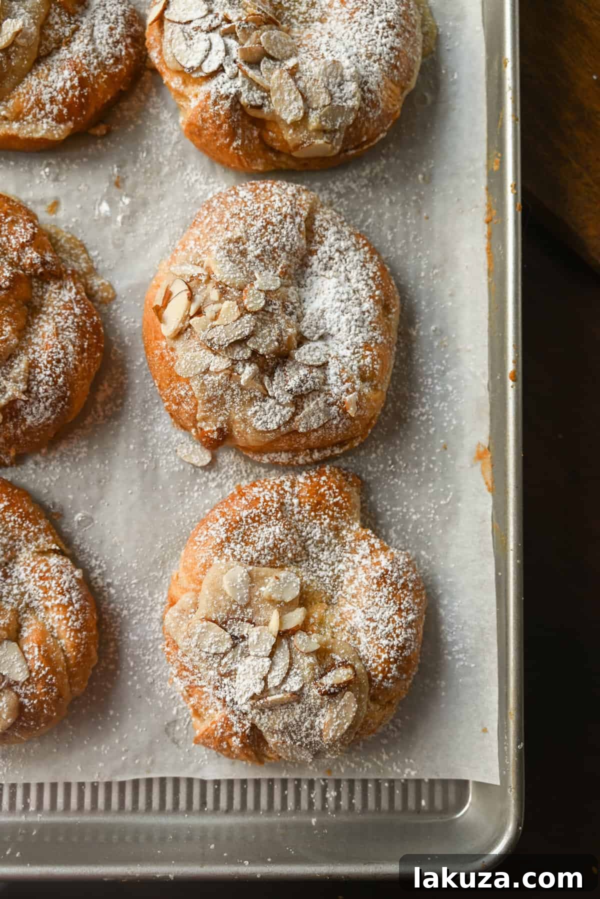 A hand dusting warm almond croissants with powdered sugar from a sieve.