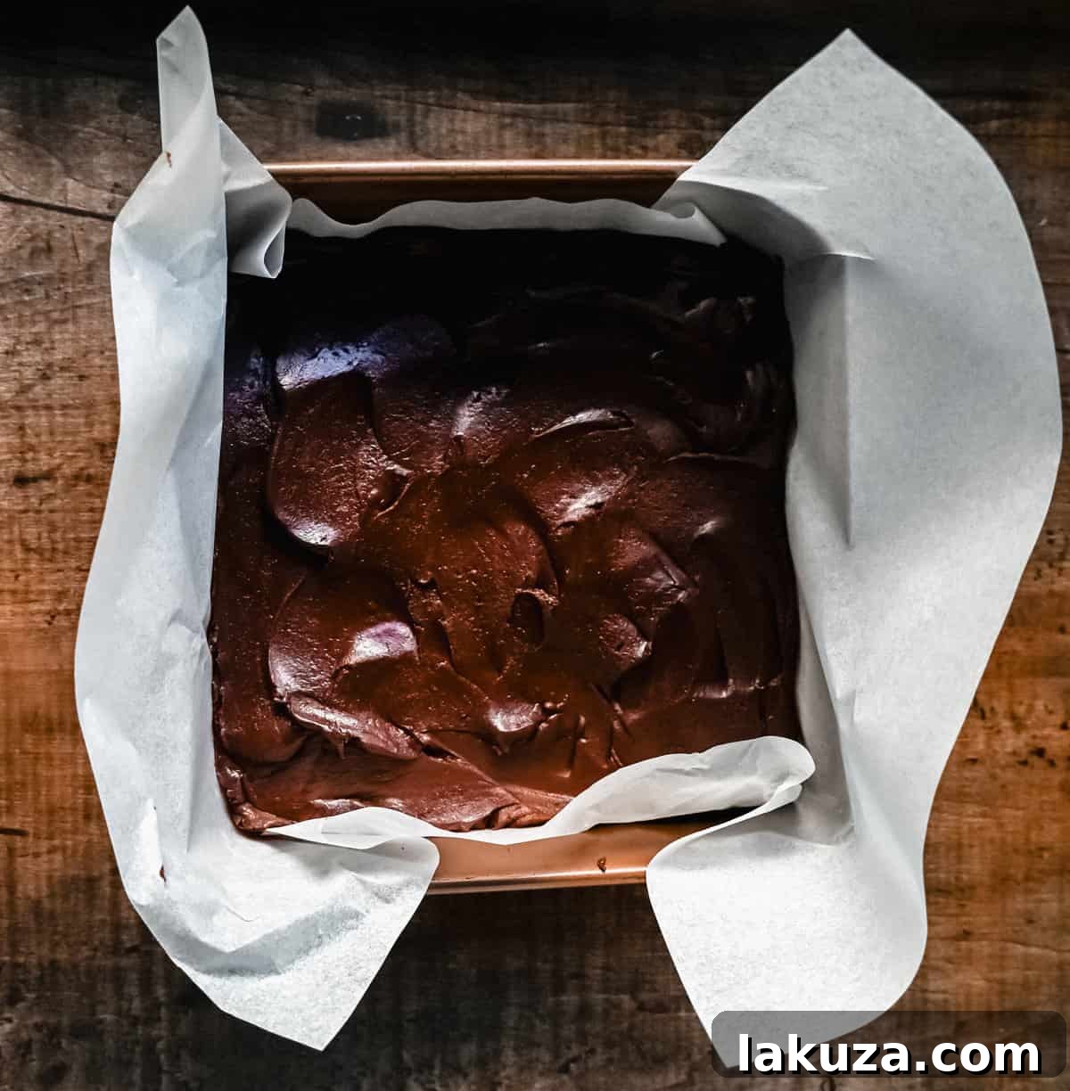 The smooth chocolate fudge mixture is poured into a parchment paper-lined baking pan, ready to be spread evenly and set.