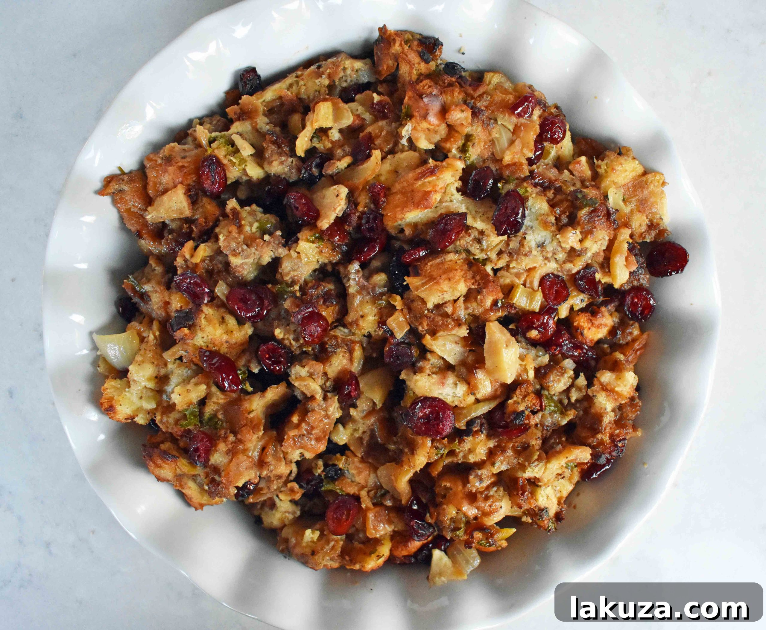 A large serving spoon scooping a generous portion of warm Cranberry Apple Sausage Stuffing from a white baking dish.