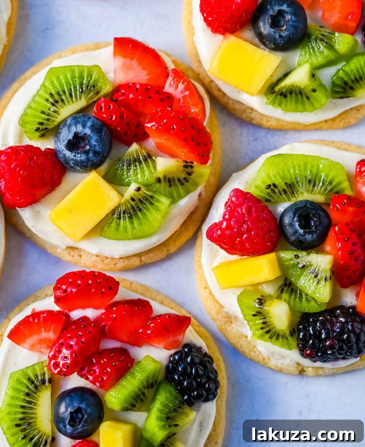 Two rows of colorful Fruit Pizza Cookies with various fresh fruits like kiwi, mango, strawberries, and blueberries on a white baking sheet.