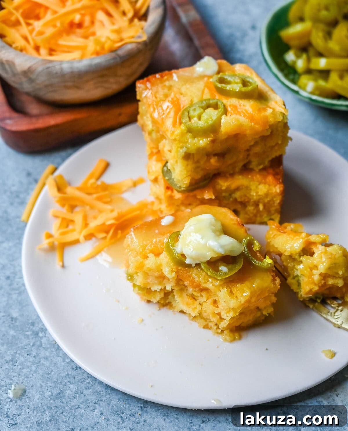 Individual ingredients for homemade jalapeno cheddar cornbread laid out on a wooden surface, including cornmeal, flour, sugar, eggs, melted butter, milk, honey, diced pickled jalapenos, and a bowl of freshly grated sharp cheddar cheese.