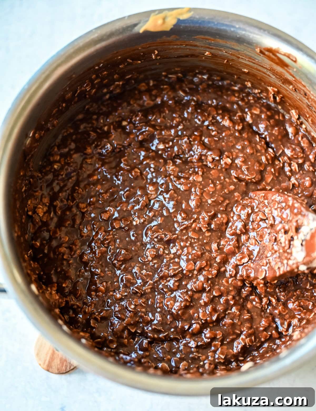 A stainless steel saucepan filled with the molten chocolate and peanut butter mixture, being stirred with a wooden spoon, showcasing the rich, glossy texture before oats are added.