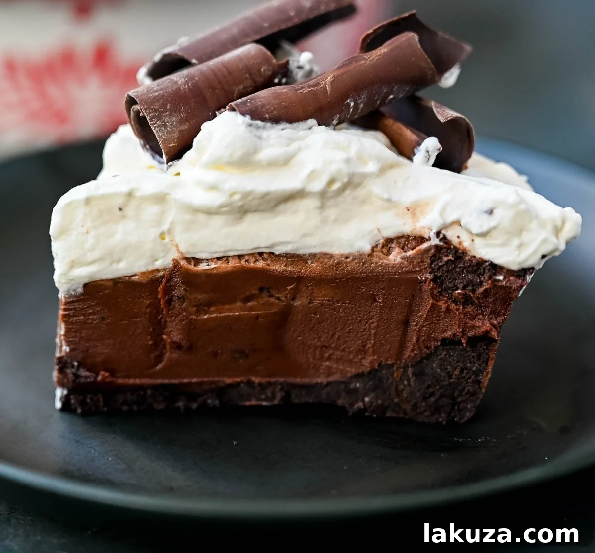 A slice of No Bake Chocolate Cream Pie revealing its layers: a dark Oreo crust, a thick, glossy chocolate ganache, and a generous topping of white whipped cream garnished with chocolate curls.