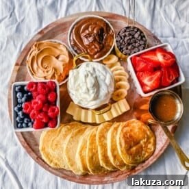 A Breakfast Pancake Board featuring homemade buttermilk pancakes and a variety of toppings like fresh berries, whipped cream, homemade syrup, Nutella, chocolate chips, and peanut butter, perfect for entertaining.