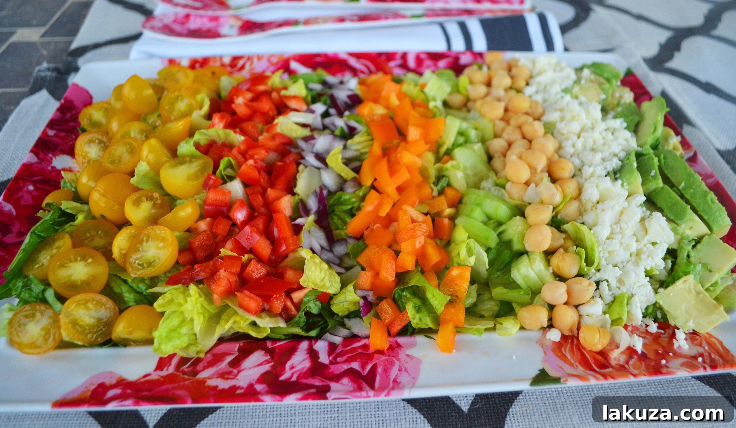An overhead view of a colorful salad bowl filled with chopped romaine, mini peppers, chickpeas, and other fresh Greek salad ingredients before being tossed.