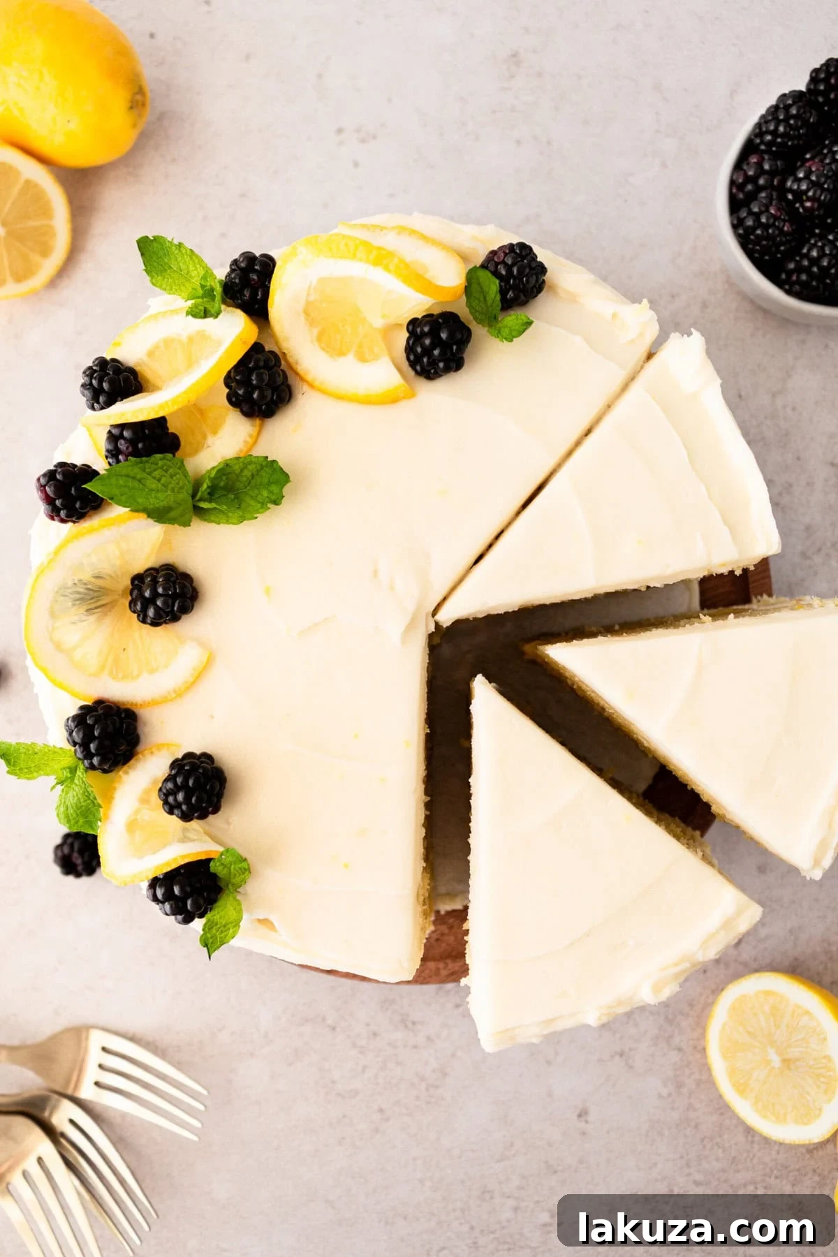 A beautifully frosted slice of Lemon Olive Oil Cake with cream cheese frosting, garnished with fresh lemon zest and blackberries, on a white plate.