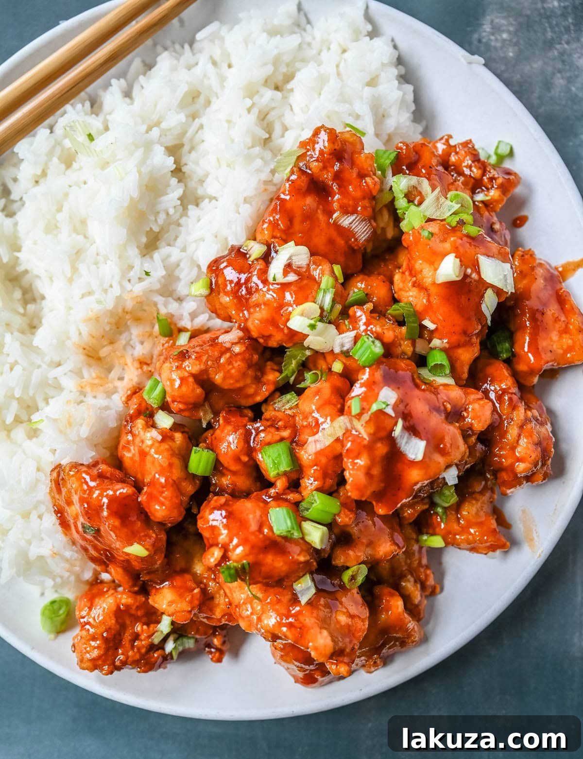 Sticky Honey Sriracha Chicken 5 A serving of homemade Honey Sriracha Chicken garnished with green onions and sesame seeds, placed next to a bowl of white rice.