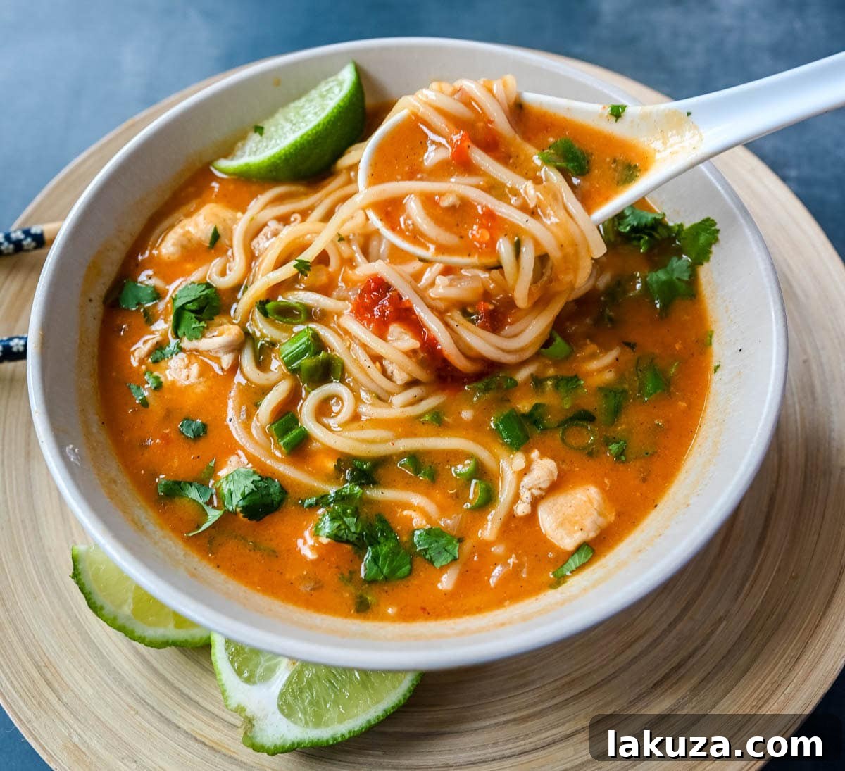 Ingredients for Chicken Curry Noodle Soup laid out on a wooden cutting board: chicken pieces, diced onion, minced garlic, grated ginger, red curry paste, chili garlic sauce, coconut milk, chicken broth, noodles, lime, and cilantro.