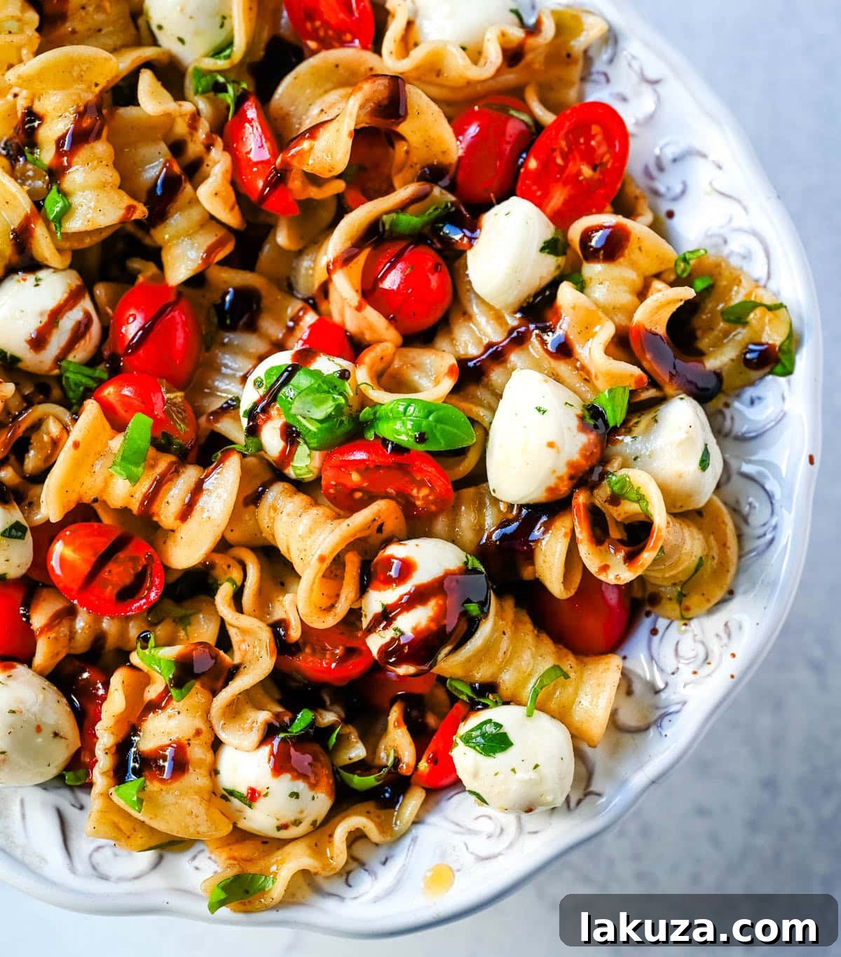 Ingredients for Caprese pasta salad laid out on a table: pasta, tomatoes, mozzarella, basil, olive oil, and balsamic vinegar.